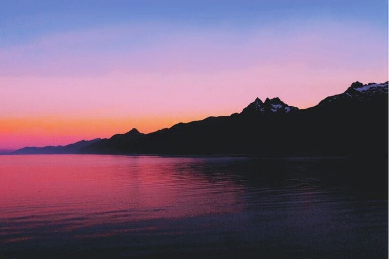freezydreamin-LXw-Dg0H4is-unsplash - Suzanne Kane A serene sunset casts vibrant colors over mountains, reflecting beautifully on the calm water below.